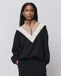 A woman wears THE GREAT. H25 TEDDY FLEECE The Colorblock Plush Chalet Sweatshirt in Almost Black, featuring a beige and white V-shaped design and half-zip collar, standing with hands in pockets against a plain background.