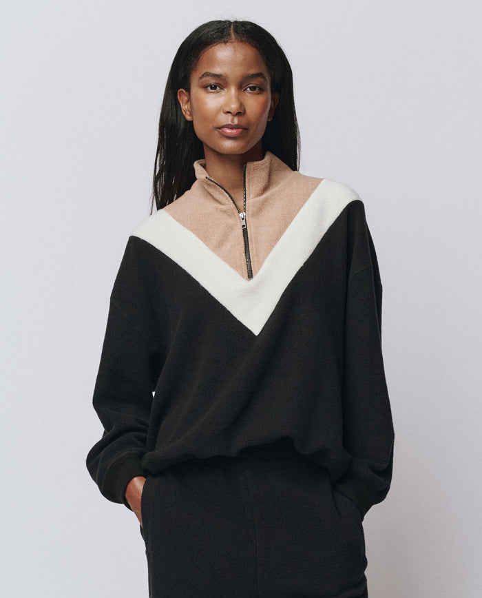 A woman wears THE GREAT. H25 TEDDY FLEECE The Colorblock Plush Chalet Sweatshirt in Almost Black, featuring a beige and white V-shaped design and half-zip collar, standing with hands in pockets against a plain background.