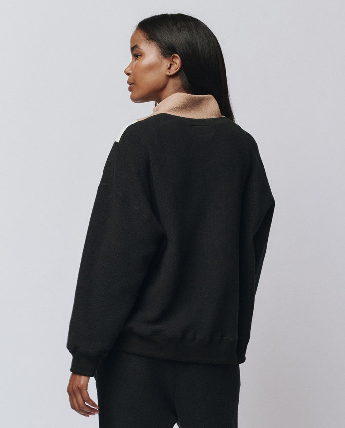 A woman with long dark hair faces away, wearing THE GREAT.’s The Colorblock Plush Chalet Sweatshirt in “Almost Black” with a beige collar, paired with matching black pants, against a plain light background.