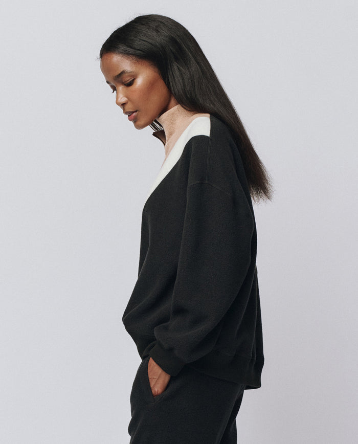 A woman stands in profile against a plain background, wearing THE GREAT. H25 TEDDY FLEECE The Colorblock Plush Chalet Sweatshirt in Almost Black with white trim and matching black pants, her hands casually tucked in her pockets.