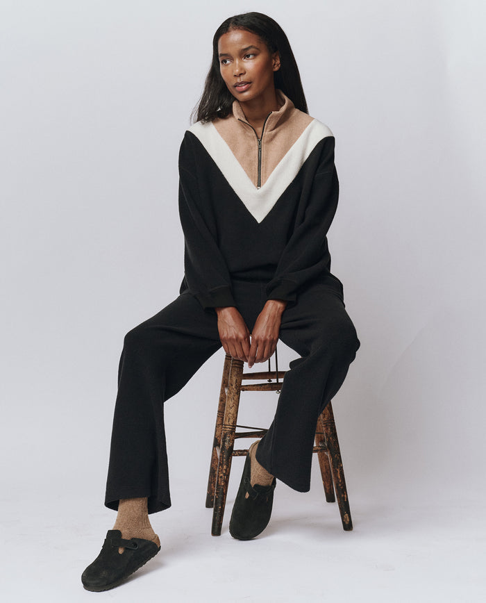 A person sits on a wooden stool wearing THE GREAT. H25 TEDDY FLEECE The Colorblock Plush Chalet Sweatshirt in Almost Black, paired with black pants, tan socks, and black slip-on shoes against a plain white background.