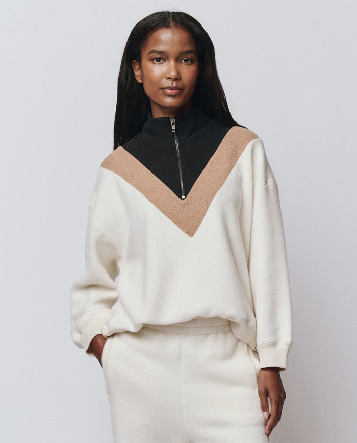 A woman stands facing forward, wearing THE GREAT. H25 TEDDY FLEECE The Colorblock Plush Chalet Sweatshirt in Washed White, styled in a relaxed fit with a black and brown V-shaped design and zipper, paired with matching cream pants.