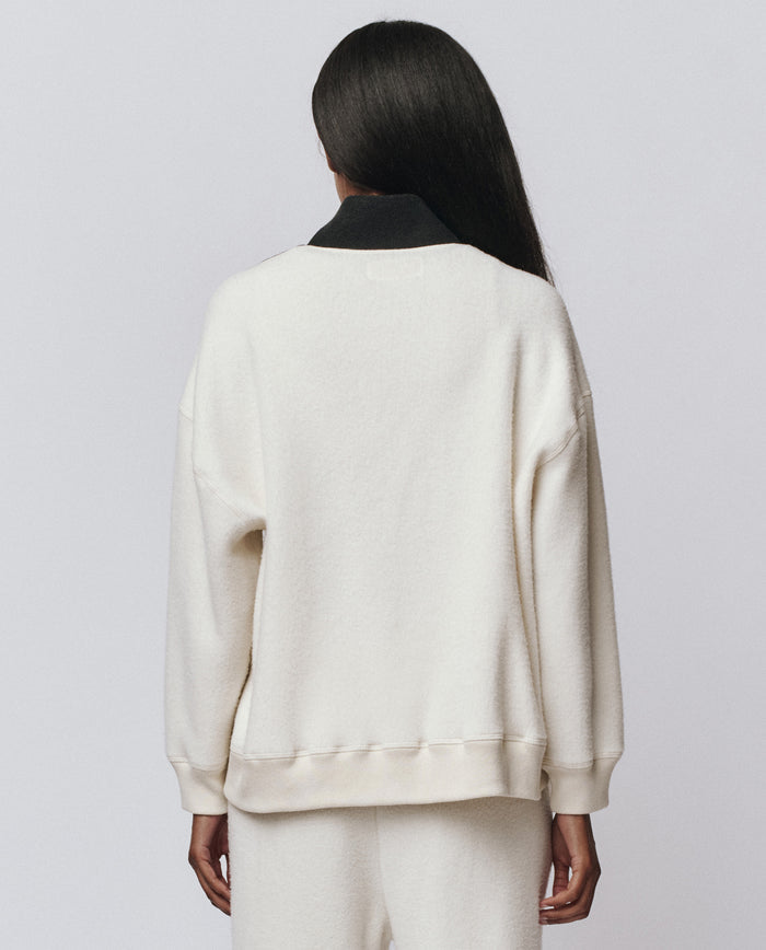 A person with long straight hair stands with their back to the camera, wearing THE GREAT. H25 TEDDY FLEECE The Colorblock Plush Chalet Sweatshirt in Washed White, paired with matching pants, against a plain light background.
