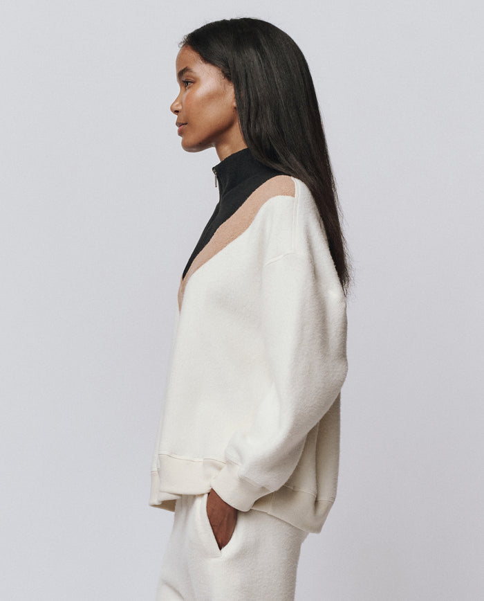 A woman stands in profile wearing THE GREAT. H25 TEDDY FLEECE Colorblock Plush Chalet Sweatshirt in Washed White with beige and black accents and white pants, all featuring a slouchy, ultra-soft fleece silhouette against a plain background.