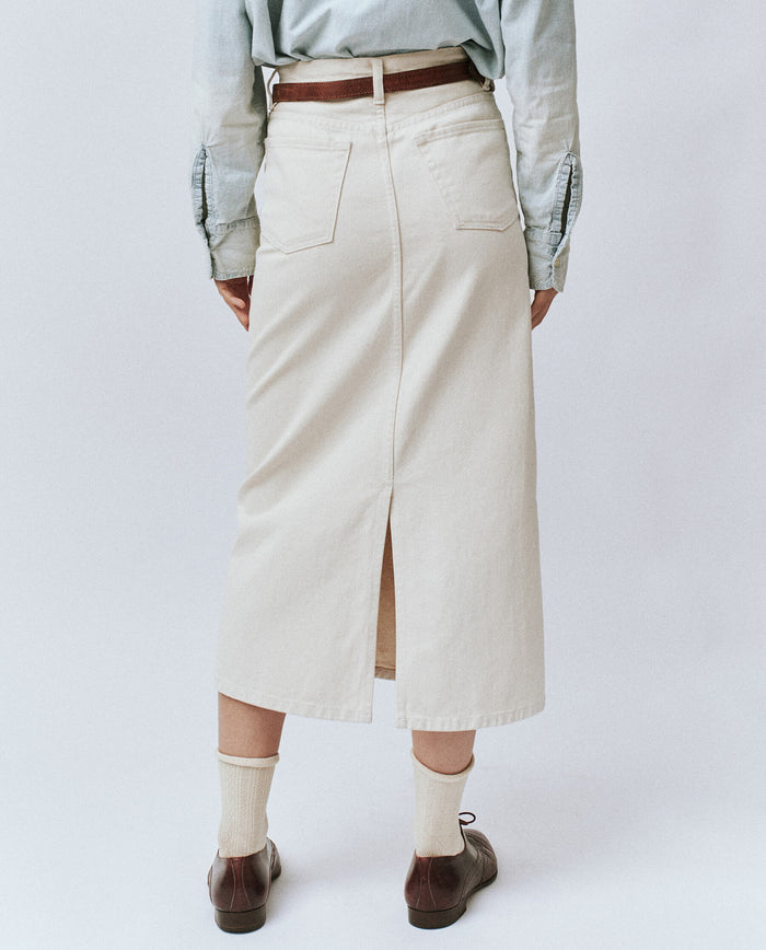 A person wearing a light blue long-sleeve shirt and The Column Skirt in Natural from THE GREAT. PF24 stands facing away, showcasing the back of the 100% cotton skirt with two pockets and a slit. They have beige socks and brown shoes on.