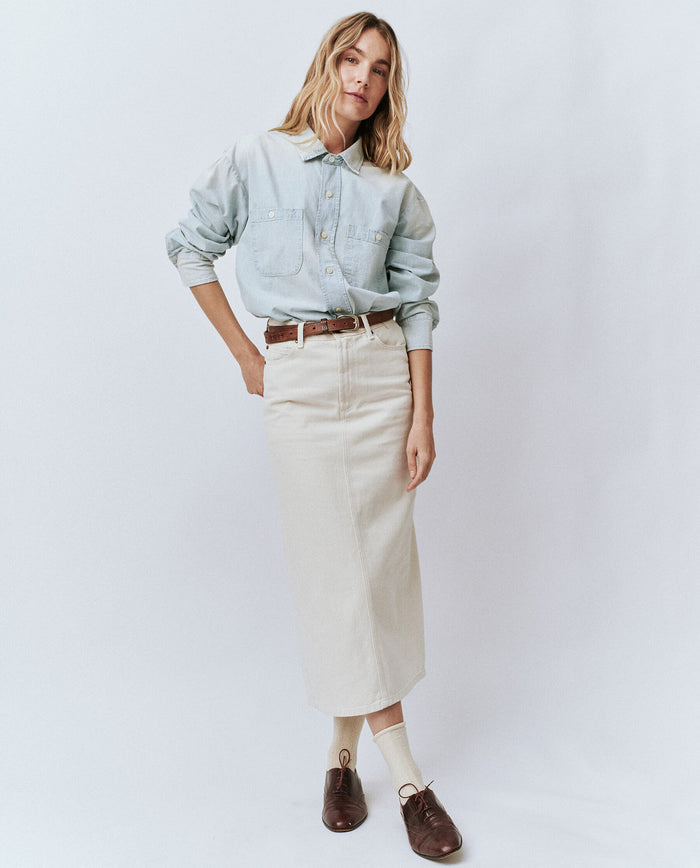A woman stands against a plain backdrop, wearing a light blue button-up shirt tucked into "The Column Skirt" in natural by THE GREAT. PF24. She has a brown belt, white socks, and brown shoes. Her hair is loosely styled, embodying a classic silhouette with timeless elegance.