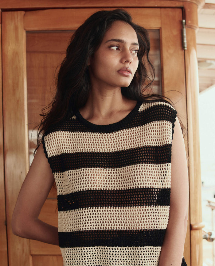 A woman with long dark hair wears THE GREAT. SU26's The Crochet Sweater Shell in Black and Cream, standing indoors before wooden doors and looking to her left.
