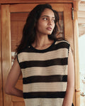 A woman with long dark hair wears THE GREAT. SU26's The Crochet Sweater Shell in Black and Cream, standing indoors before wooden doors and looking to her left.