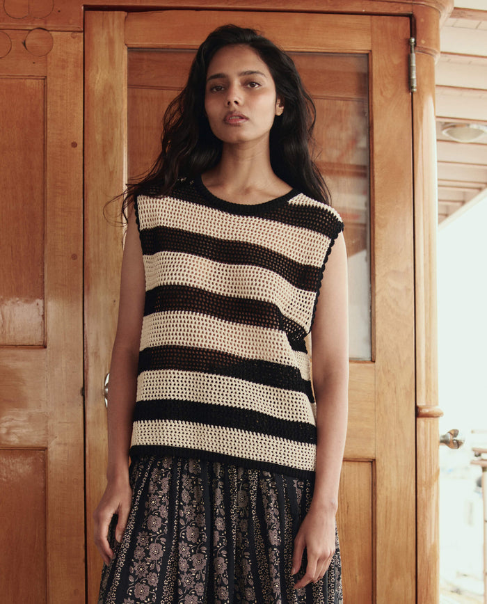 A woman stands before a wooden door, wearing THE GREAT. SU26's The Crochet Sweater Shell in Black and Cream paired with a patterned skirt.