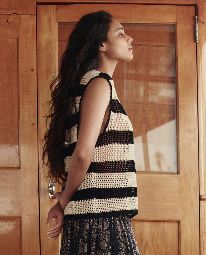 A woman with long dark hair stands in profile wearing THE GREAT. SU26's The Crochet Sweater Shell in Black and Cream, paired with a patterned skirt, in front of a wooden door.
