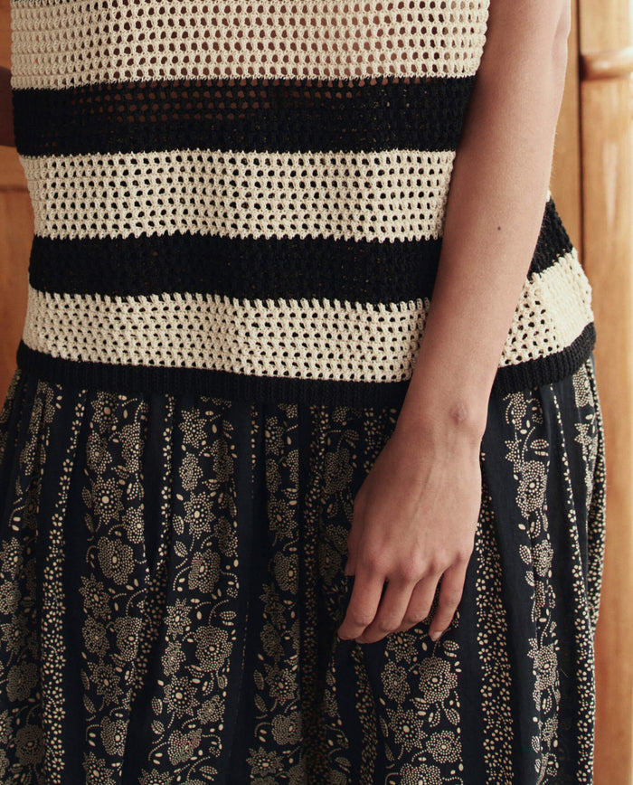 A person wearing a sleeveless black and white striped crochet top and THE GREAT. SU26 The Canary Skirt in Black And Cream Token Floral, standing indoors.