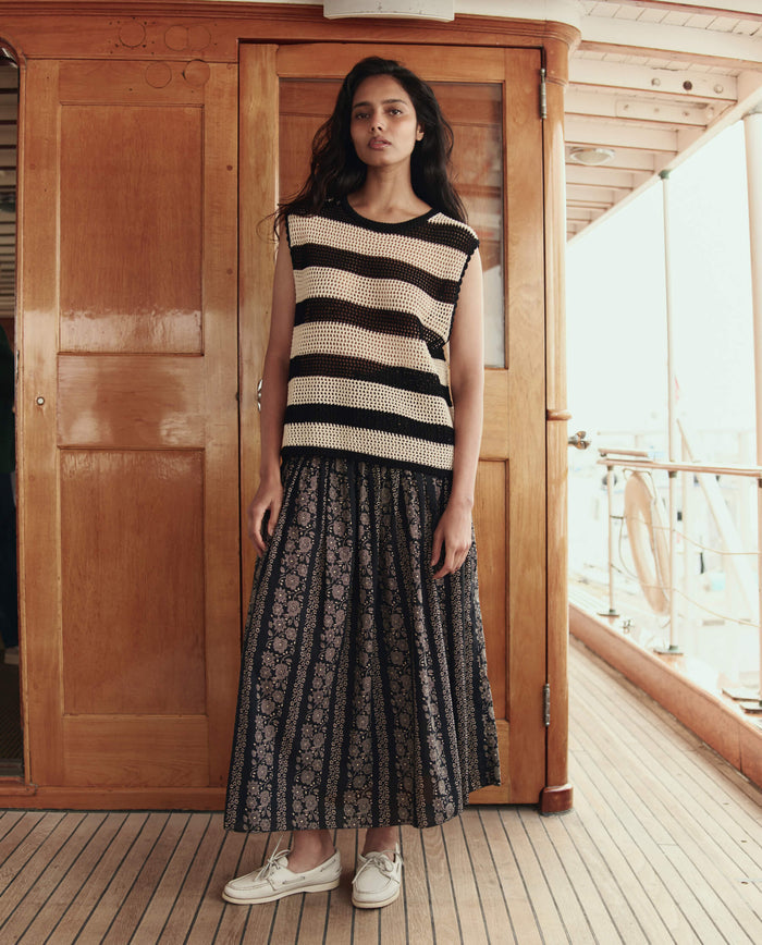 A woman stands on a wooden ship deck, wearing THE GREAT. SU26 The Crochet Sweater Shell in black and cream, paired with a patterned long skirt and beige shoes.