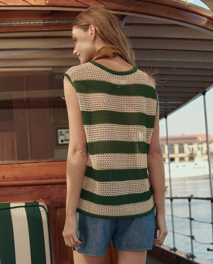 A woman with long hair stands on a wooden boat, wearing THE GREAT. SU26's Grove And Cream Crochet Sweater Shell and blue shorts, gazing toward the water.