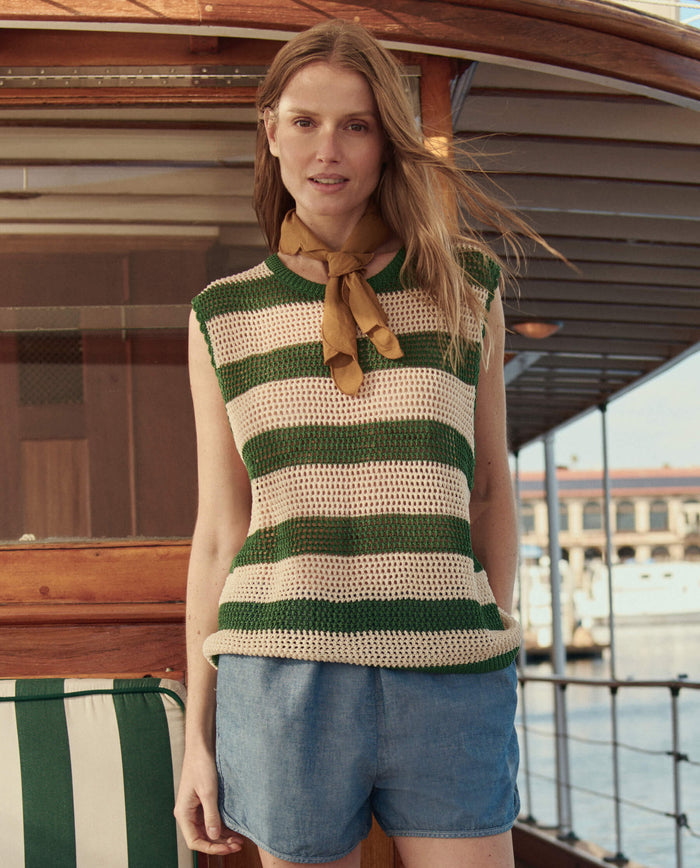 A woman stands on a boat wearing THE GREAT. SU26 The Crochet Sweater Shell in Grove and Cream, paired with denim shorts and a yellow scarf. A marina and buildings appear in the background.