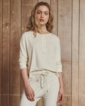 A person with long, wavy blonde hair wears THE GREAT. H25 SLEEP's The Crop Sleep Henley in Washed White, paired with matching cotton drawstring pants, standing before a wooden plank wall.