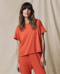 A woman with long brown hair stands against a beige fabric backdrop, modeling THE GREAT. SU25 SEASONAL KNITS’ The Crop Tee. Solid -- Desert Clay, paired with matching pants for a stylish, casual look in women’s fashion.