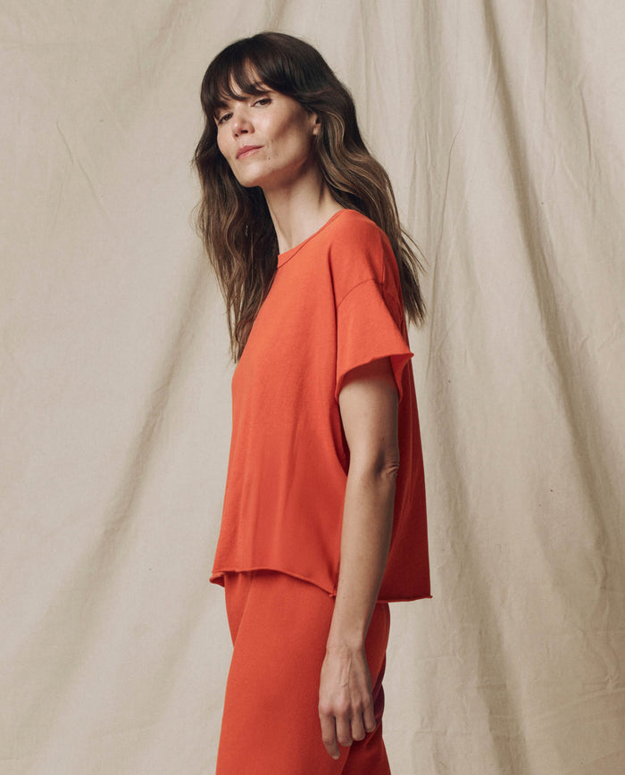 A woman with long brown hair models THE GREAT. SU25 SEASONAL KNITS The Crop Tee. Solid in Desert Clay, paired with matching pants against a beige fabric backdrop, exemplifying easy women's fashion.