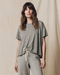 A person in THE GREAT. CORE KNITS' Heather Grey Crop Tee and matching drawstring pants stands against a beige fabric backdrop.
