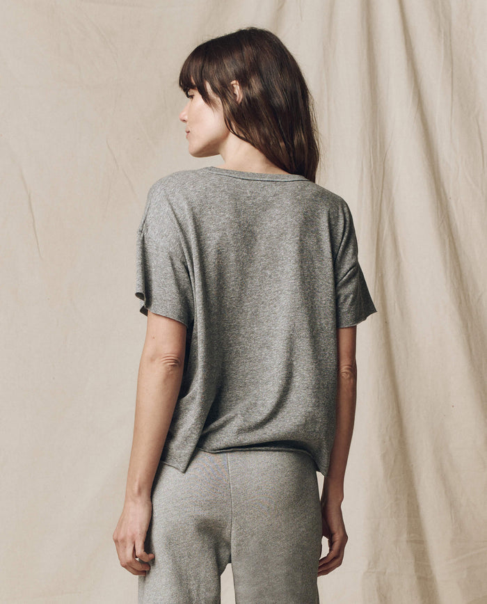 A person with long hair stands sideways, wearing The Crop Tee in Heather Grey by THE GREAT. CORE KNITS, made of super soft cotton, along with pants. The background is plain beige fabric.