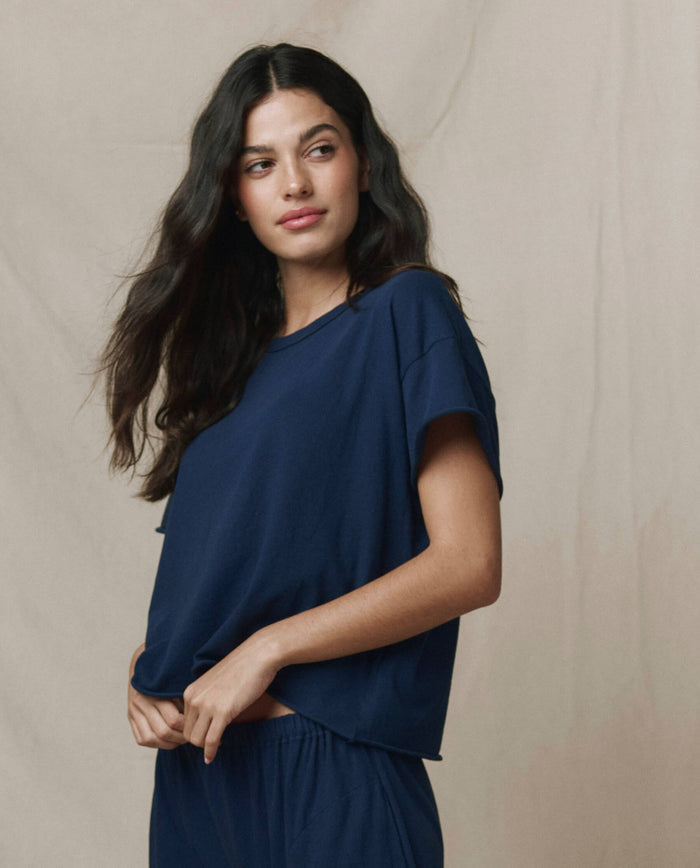 A woman with long dark hair wears THE GREAT. CORE KNITS The Crop Tee. Solid in True Navy, paired with matching pants, standing against a beige background.