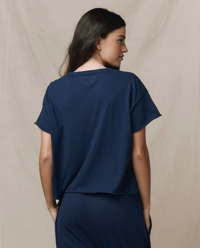 A woman with long dark hair wears THE GREAT. CORE KNITS The Crop Tee. Solid in True Navy, paired with matching pants, standing facing away from the camera against a beige backdrop.