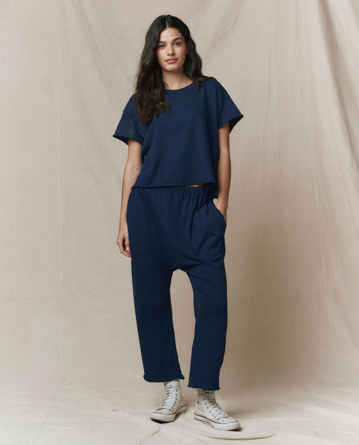 A person stands against a neutral backdrop wearing THE GREAT. CORE KNITS The Crop Tee. Solid in True Navy, styled as a loose-fitting matching set with short sleeves and paired with white high-top sneakers.