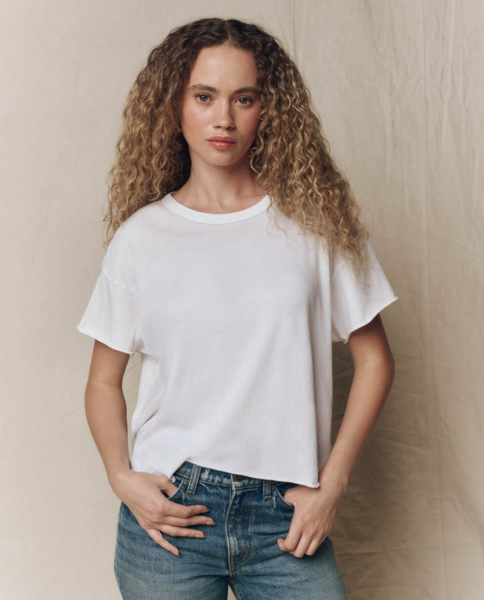 A woman with curly hair wears THE GREAT. CORE KNITS The Crop Tee. Solid in True White and blue jeans, standing before a beige backdrop and looking at the camera with a neutral expression.