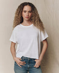 A woman with curly hair wears THE GREAT. CORE KNITS The Crop Tee. Solid in True White and blue jeans, standing before a beige backdrop and looking at the camera with a neutral expression.