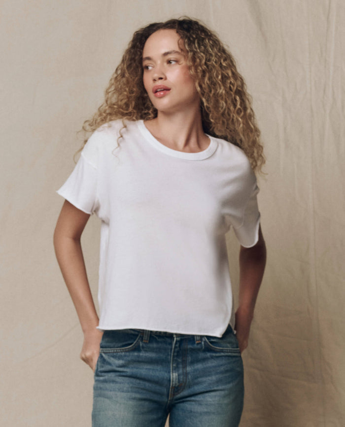 A person with curly hair wears THE GREAT. CORE KNITS The Crop Tee. Solid -- True White and blue jeans, standing in front of a neutral, textured background.