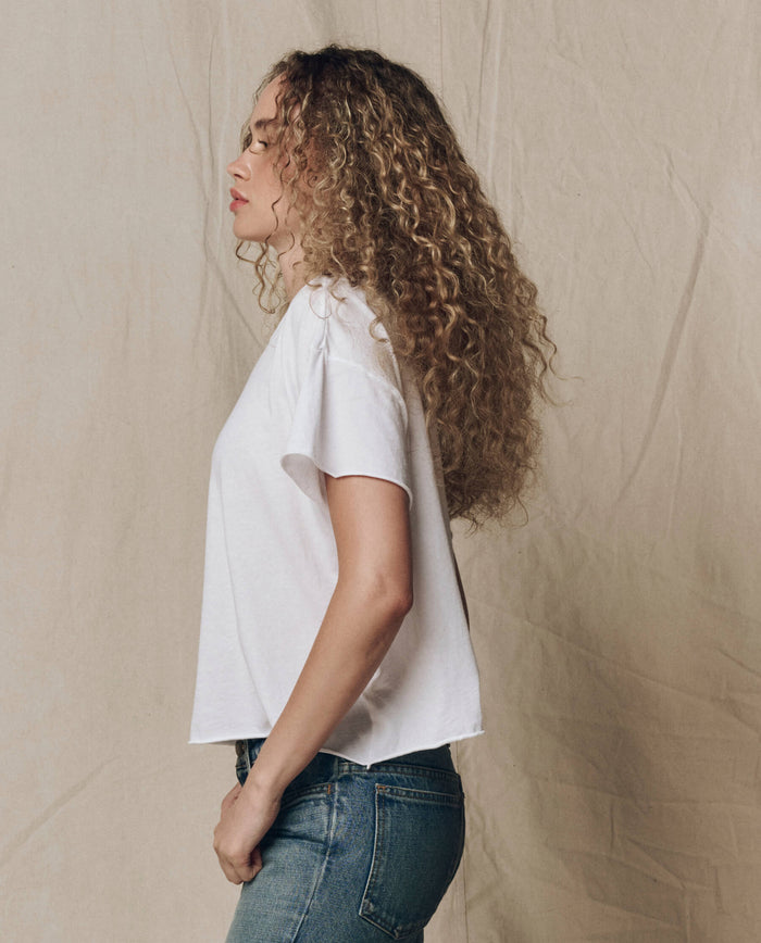 A person with long curly hair stands in profile, wearing THE GREAT. CORE KNITS The Crop Tee. Solid -- True White and blue jeans, set against a beige fabric background.