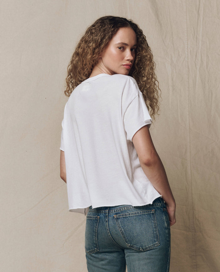Someone with curly hair wears THE GREAT. CORE KNITS The Crop Tee. Solid -- True White and blue jeans, standing sideways and turning slightly toward the camera against a neutral fabric backdrop.