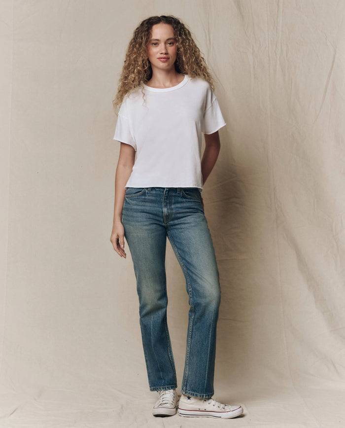 A person with curly hair stands against a beige backdrop, wearing THE GREAT. CORE KNITS The Crop Tee. Solid -- True White, blue jeans, and white sneakers.