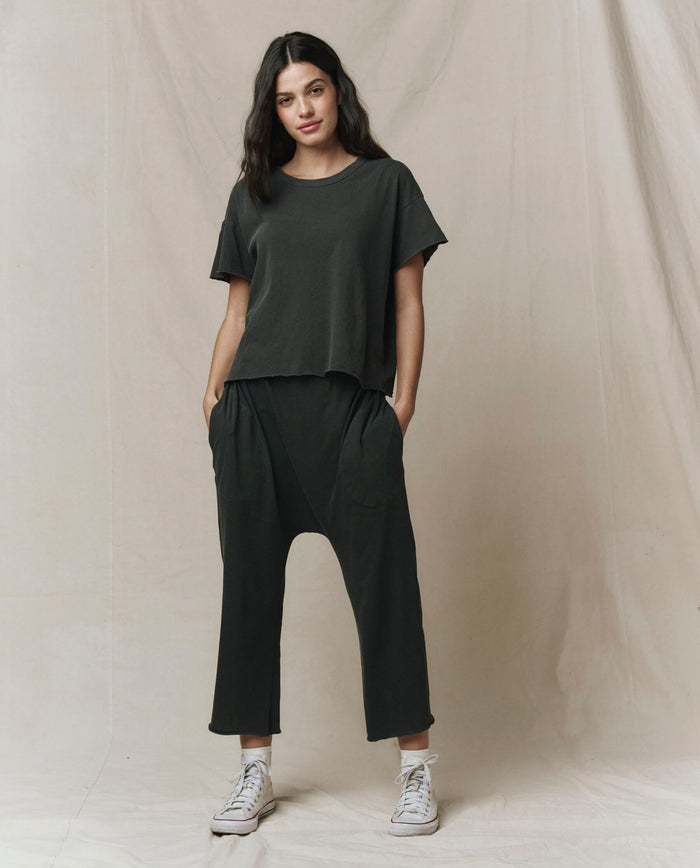 A woman stands against a neutral background wearing THE GREAT. CORE KNITS The Crop Tee. Solid -- Washed Black, styled with black drop-crotch pants and white sneakers, hands in her pockets. Super soft cotton. Made in USA.
