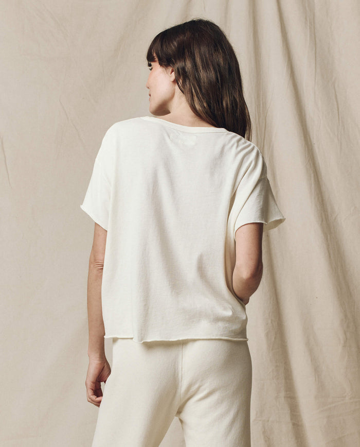 A person with long brown hair stands in front of a beige fabric backdrop, wearing THE GREAT. CORE KNITS' The Crop Tee in Washed White and super soft cotton pants.