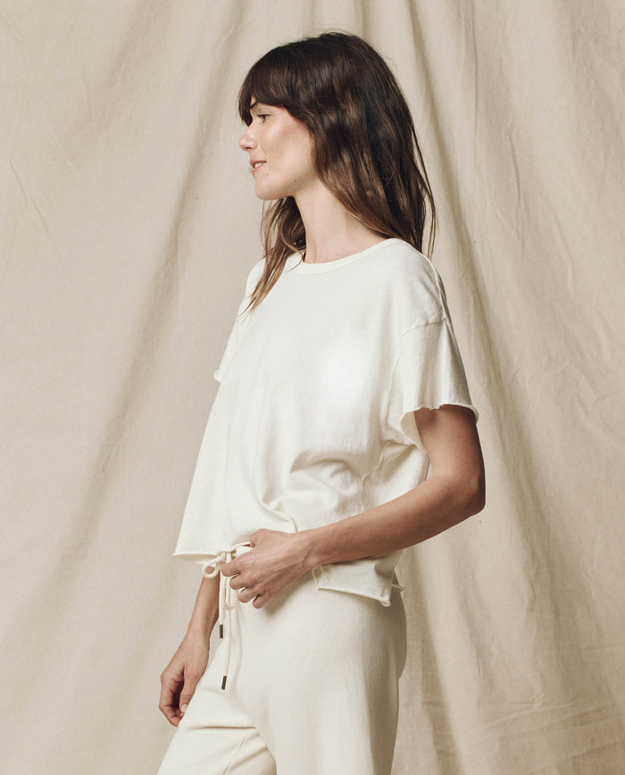 A person wearing "The Crop Tee. Solid -- Washed White" by THE GREAT. CORE KNITS stands against a draped fabric background, looking to the side.