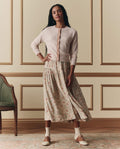 A woman stands indoors, wearing EMME's The Dainty Cardigan in Soft Petal, paired with a long floral skirt, cream socks, and brown shoes. One foot is slightly raised beside a green chair.