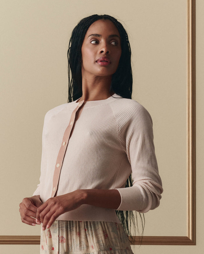 A woman with long braided hair models EMME’s The Dainty Cardigan in Soft Petal and a floral skirt, standing before a beige background with gold framing.