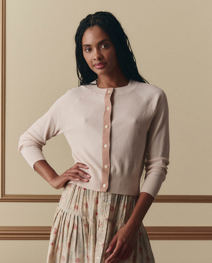 A woman with braided hair wears EMME's The Dainty Cardigan in Soft Petal, paired with a patterned skirt, standing against a beige paneled background.