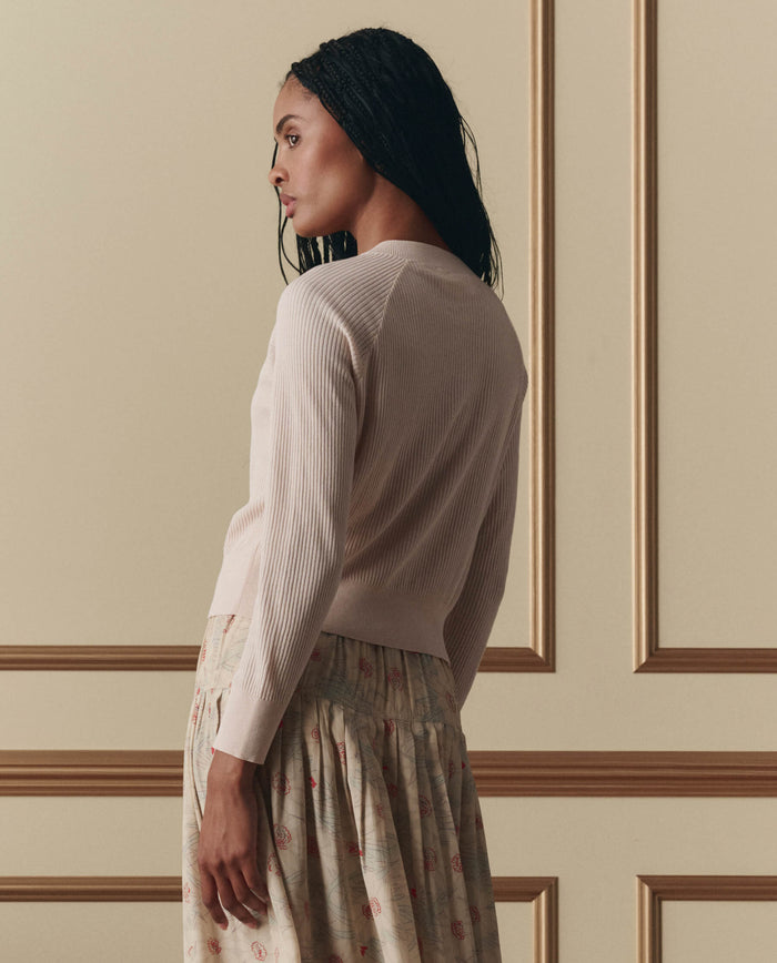 A woman with braided hair stands sideways in front of a beige wall with decorative molding, wearing EMME’s The Dainty Cardigan in Soft Petal and a patterned skirt.