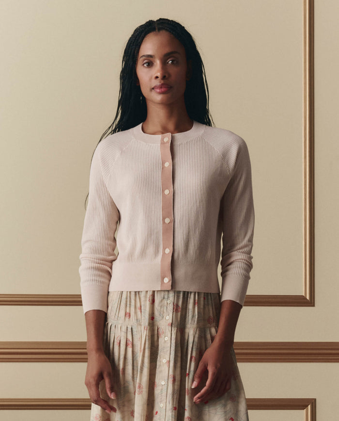 A woman stands indoors against a beige wall with molding, wearing EMME’s The Dainty Cardigan in Soft Petal and a patterned skirt.
