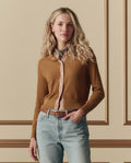 A woman with long blonde hair wears THE GREAT. SP26 D2’s The Dainty Cardigan in Suntan over a pink shirt, blue jeans, and a belt, standing against a beige wall with decorative molding.