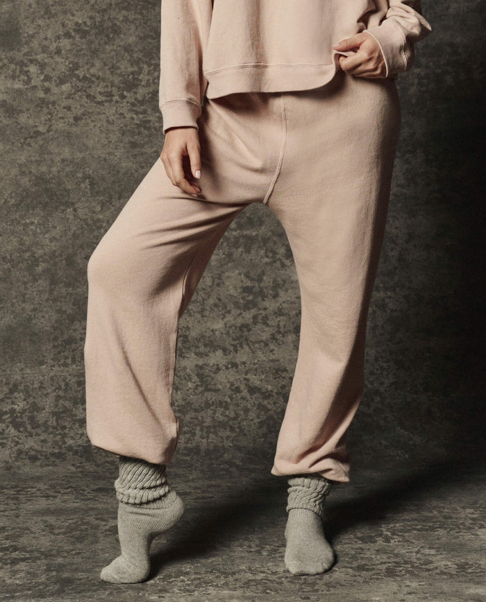 Wearing THE GREAT. PF26 DANCER KNIT BODIES' The Dance Pant in Ballet Pink, a person pairs the look with a matching sweatshirt and gray socks, standing on a textured surface with one foot flat and the other tiptoe—an effortless showcase of relaxed, comfortable dancewear style.