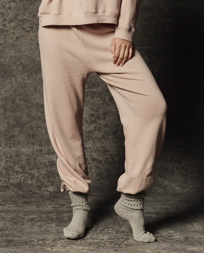 A person wearing THE GREAT. PF26 DANCER KNIT BODIES The Dance Pant in Ballet Pink and gray socks stands on a textured gray surface, one foot slightly raised and a hand by their side, highlighting these comfortable dance pants ideal for movement and relaxation.