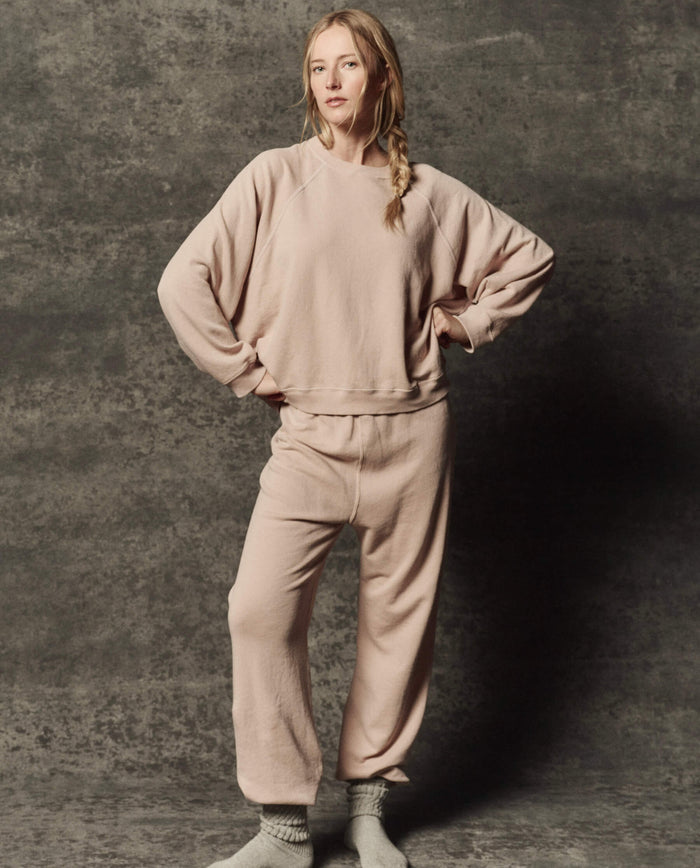 A person stands against a textured gray backdrop, wearing THE GREAT. PF26 DANCER KNIT BODIES' The Dance Pant in Ballet Pink paired with a light beige sweatshirt and gray socks, hands on hips and looking at the camera.