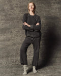 Standing against a textured gray background, the person wears THE GREAT. PF26 DANCER KNIT BODIES' The Dance Pant in Flat Charcoal, paired with a loose dark sweatshirt and light gray socks. Their arms are crossed, and they hold a neutral expression.