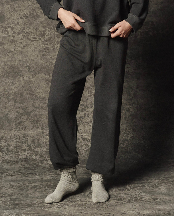 Wearing THE GREAT. PF26 DANCER KNIT BODIES The Dance Pant in Flat Charcoal with a coordinating sweatshirt and light gray socks, the individual stands on a textured surface, hands gently adjusting the waistband.