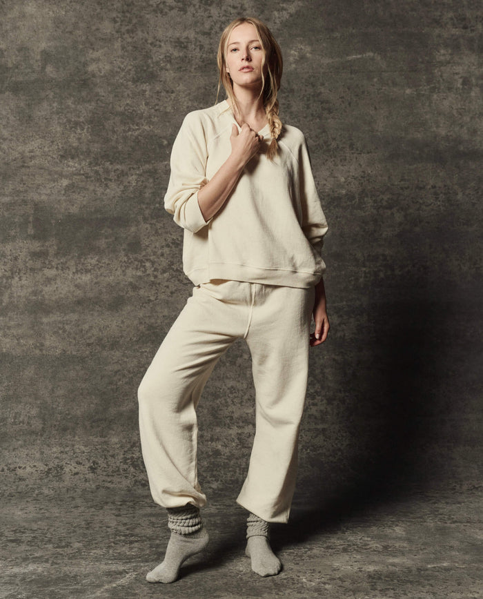 A person wearing THE GREAT. PF26 DANCER KNIT BODIES' The Dance Pant in Washed White stands against a textured dark gray backdrop with gray socks and one hand touching their collar.