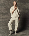 A person wearing THE GREAT. PF26 DANCER KNIT BODIES' The Dance Pant in Washed White stands against a textured dark gray backdrop with gray socks and one hand touching their collar.