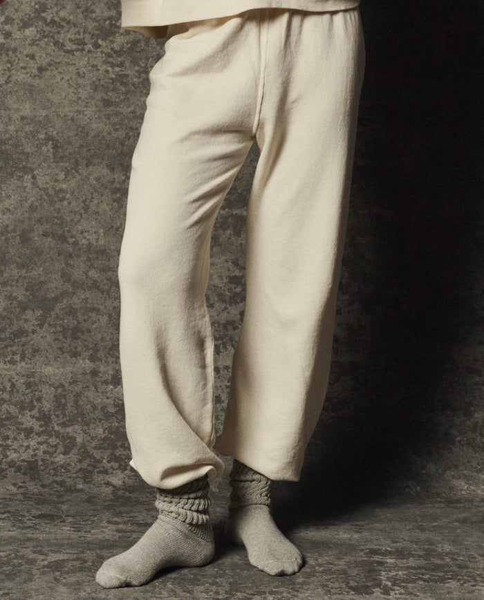 A person wearing THE GREAT. PF26 DANCER KNIT BODIES' The Dance Pant in Washed White and gray socks stands on a textured floor with a matching textured background; upper body not visible.
