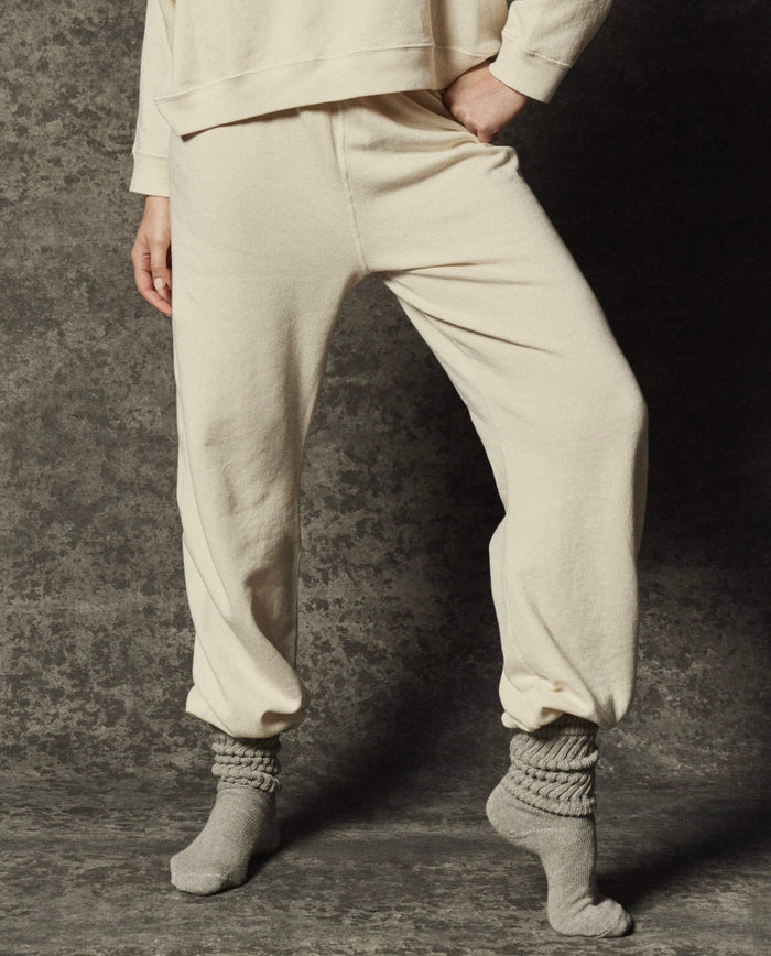 A person stands against a textured gray background wearing THE GREAT.’s The Dance Pant in Washed White from the PF26 Dancer Knit Bodies collection, paired with a matching sweatshirt and gray socks.